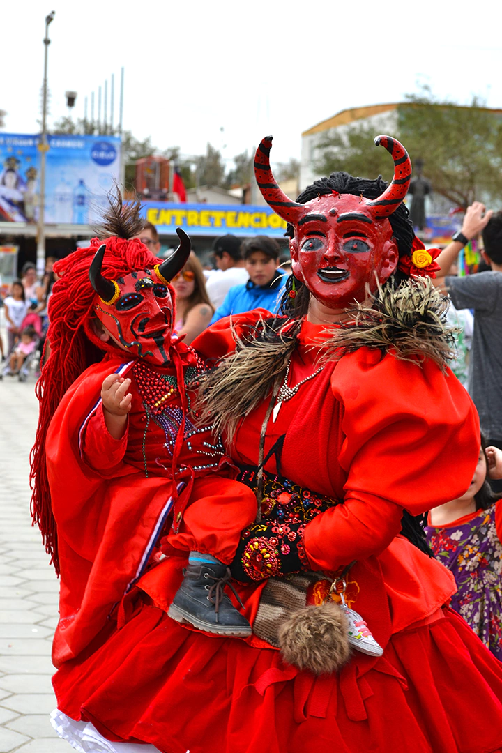 Fiesta de la tirana-diablada-madre e hija-Foto Marianne Fuentealba La mujer y su hijo están vestidos con trajes tradicionales de diablo suelto de La Tirana. Los trajes son rojos y están decorados con lentejuelas, espejos y motivos religiosos. Llevan máscaras de yeso o papel maché que representan figuras demoníacas.